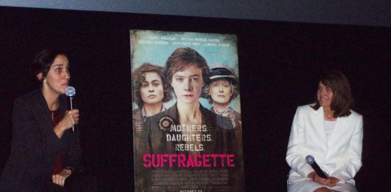 Two women sit next to poster of Suffragette film.