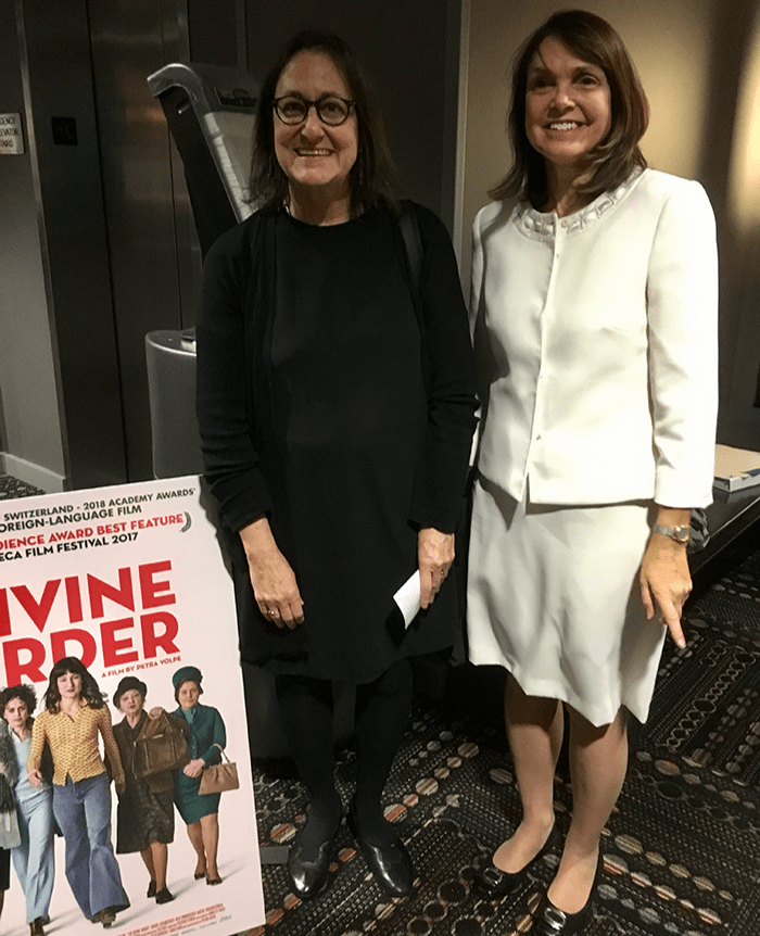 Two women stand next to movie poster for "Divine Order."