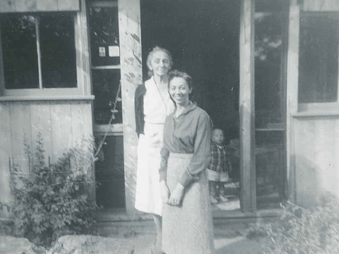 Black and white photo of young woman and older woman in front of wooden house.